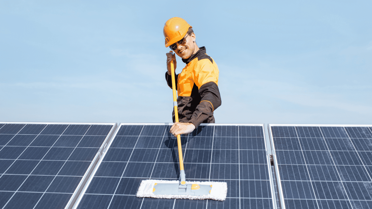 Smiling worker in a hard hat cleaning solar panels with a mop under clear sky.
